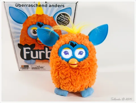 Orange Furby 2012 with blue ears and faceplate beside its original box