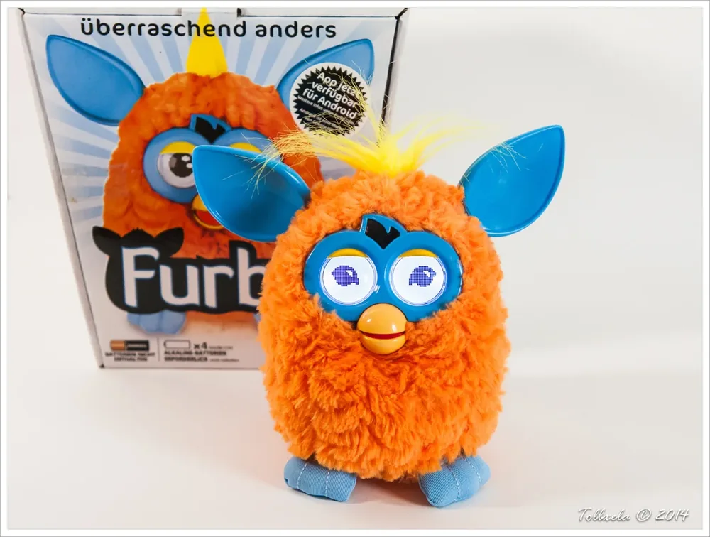 Orange Furby 2012 with blue faceplate and ears beside its product box.